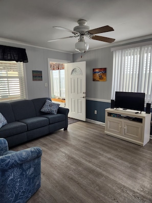 Un salon comprenant un canapé, un téléviseur posé sur un meuble, un ventilateur de plafond et une porte avec une fenêtre.