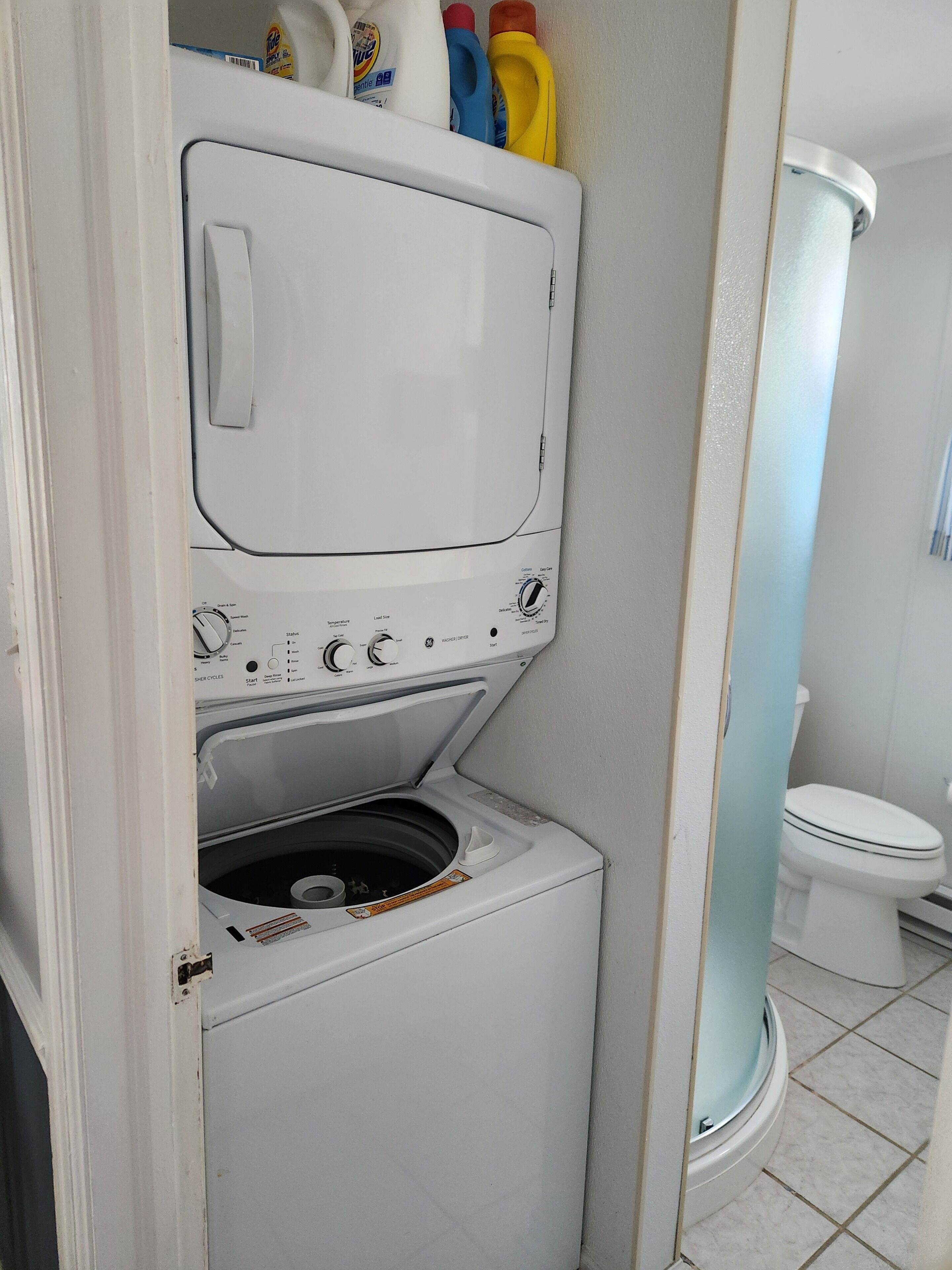 A compact, white washing machine in a bathroom with a toilet and a towel rack.