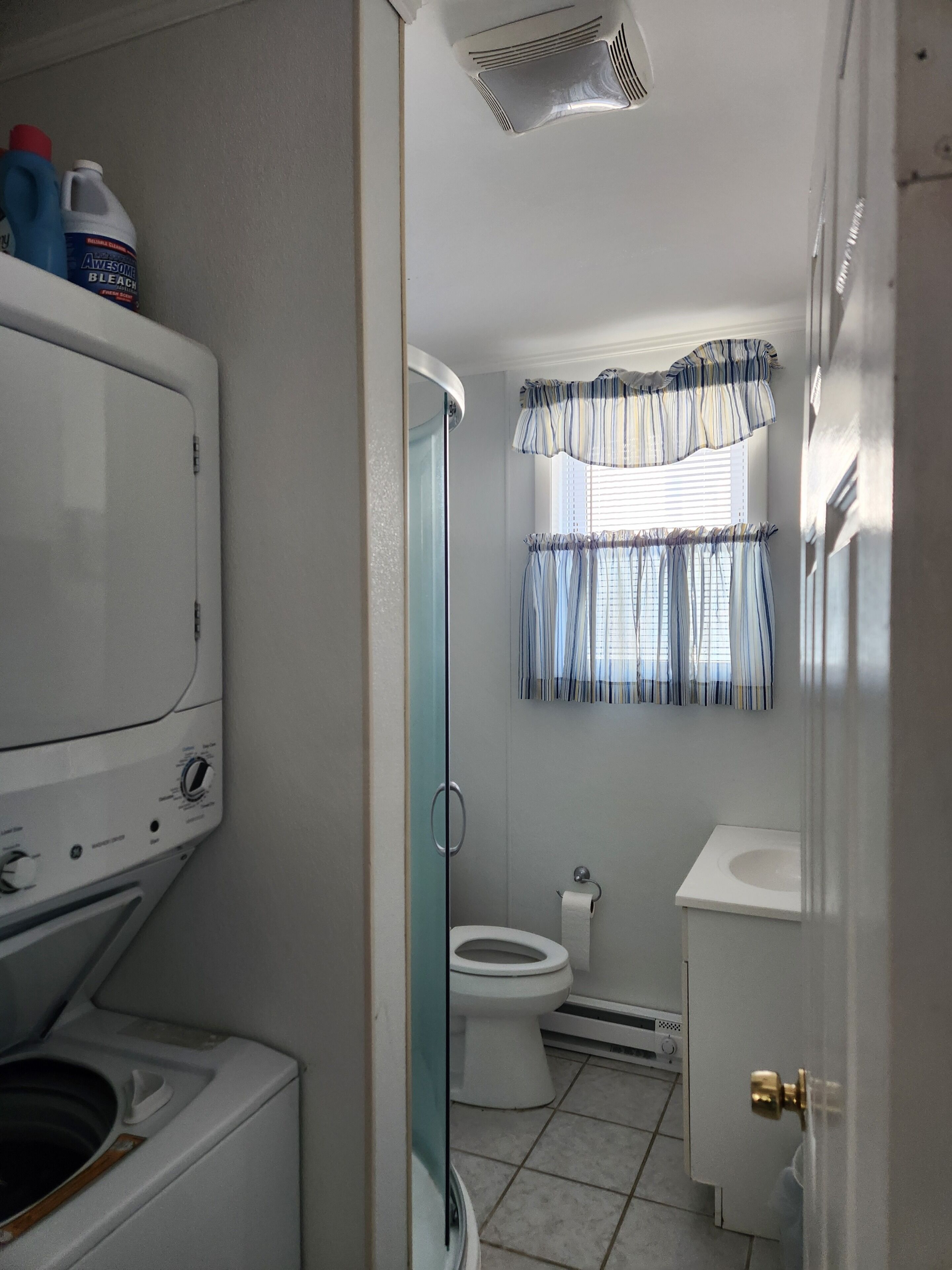 A compact bathroom with a washing machine, toilet, and a window with curtains.