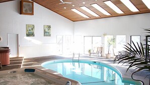 An indoor swimming pool with a wooden ceiling, a hot tub, and a seating area with chairs and potted plants.