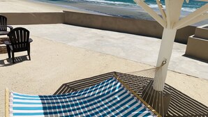 A beachside patio with a white umbrella, black chairs, and a blue-striped beach mat.