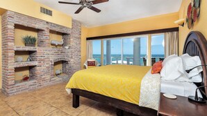 A bedroom with a bed, a brick fireplace, a view of the ocean, and a ceiling fan.