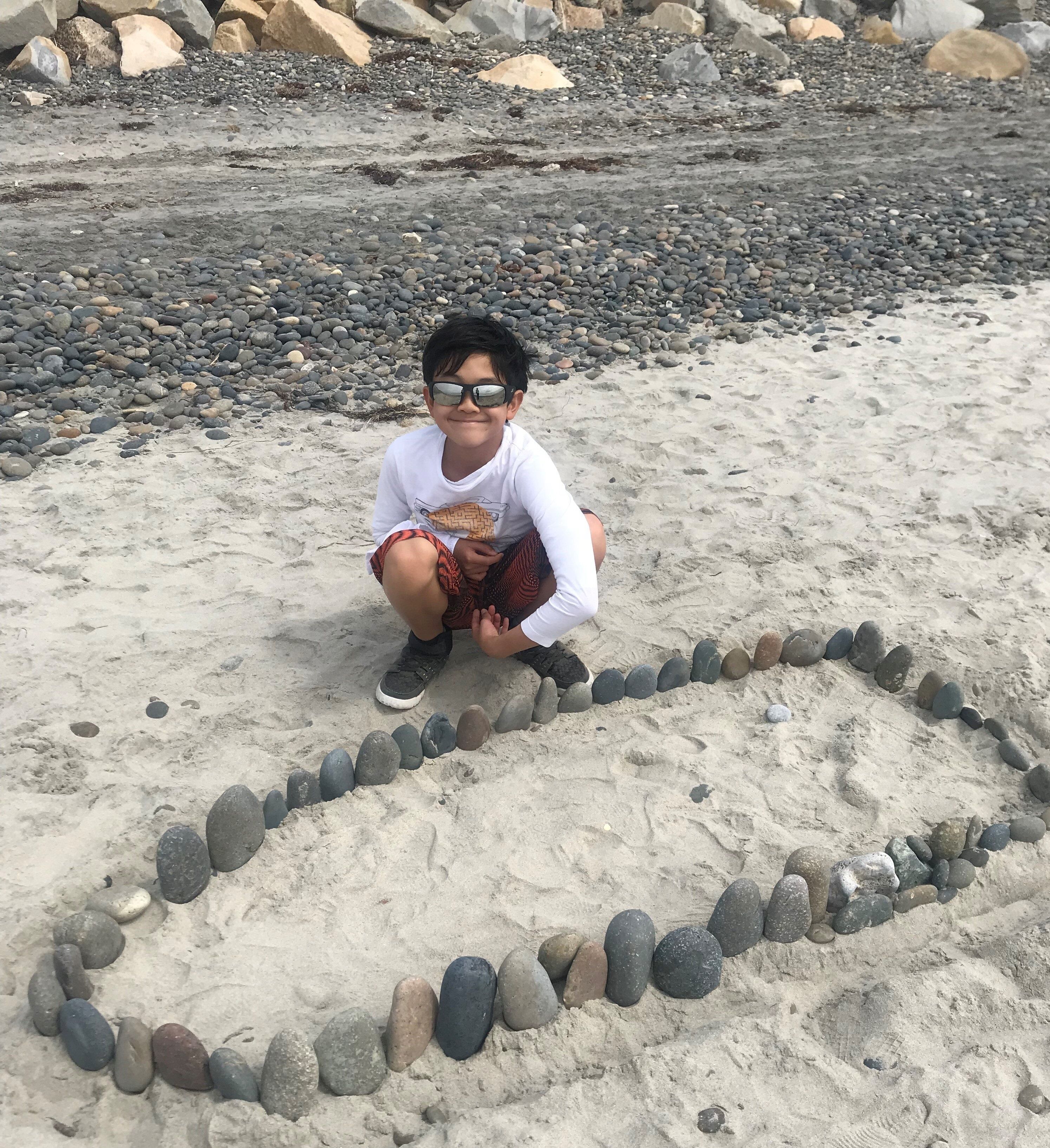 Une personne accroupie sur une plage de sable, devant une composition de pierres en forme de cœur.