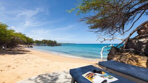 A beach with a lounge chair and a magazine, overlooking a clear blue sea and a rocky shoreline.