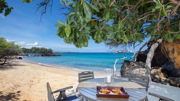 A beachside table with chairs, a tray with drinks, and a candle, overlooking a clear blue sea and sandy beach.