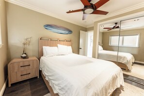 Une chambre à coucher comprenant un lit, une table de chevet, un ventilateur de plafond, une planche de surf accrochée au mur et un miroir.