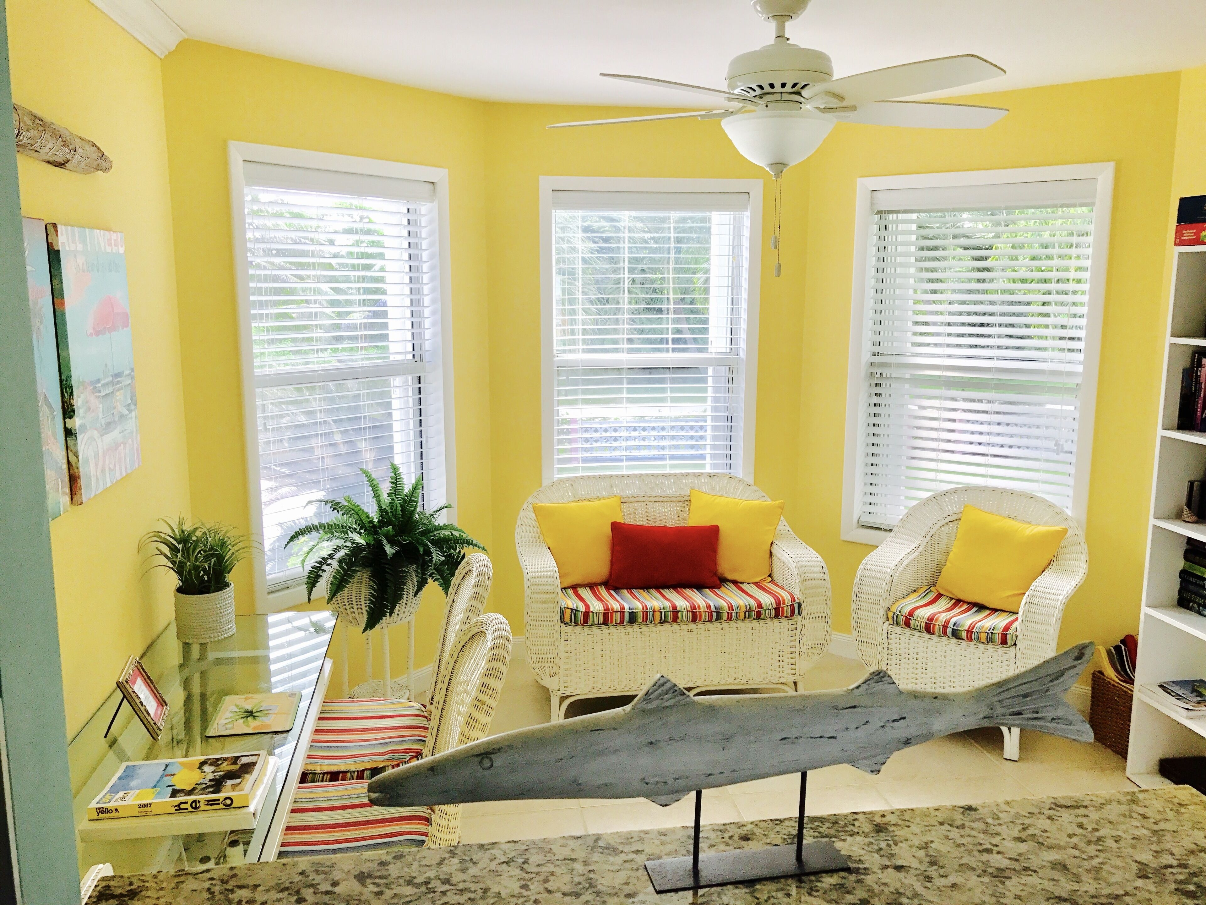 A room with yellow walls, white wicker furniture, a ceiling fan, and a large fish sculpture on the floor.