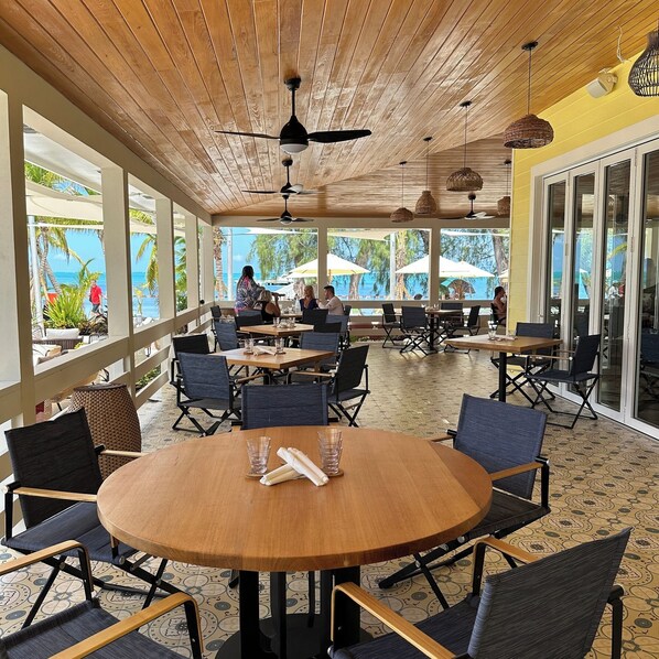 Ein Außenbereich zum Essen mit Holztischen und -stühlen, Deckenventilatoren und großen Fenstern mit Blick auf das Meer und Palmen.