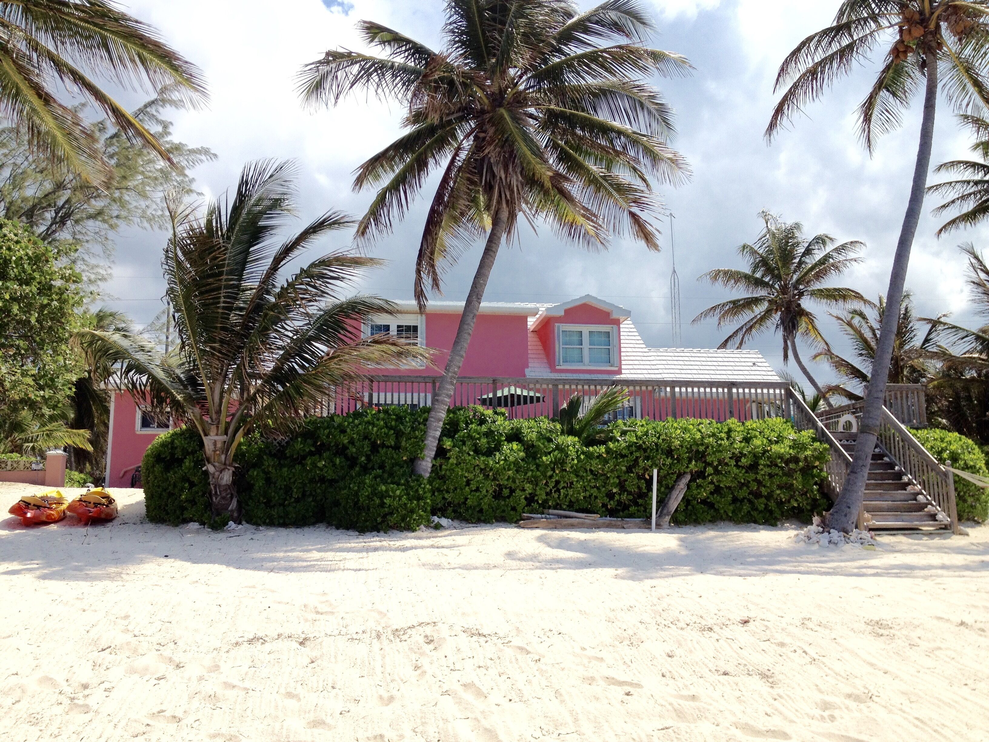 Une maison rose avec un toit blanc, entourée de palmiers et bordée d’une plage de sable.