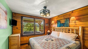 A cozy bedroom with a wooden headboard, a large window with blinds, a ceiling fan, and a quilted bedspread.