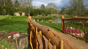 A wooden bridge over a pond with a rainbow in the background, surrounded by trees and a grassy area. - Silver Lining Cabin -Natural light, Quiet, Hot Tub, Nature Trails, Pet door (Fletcher)