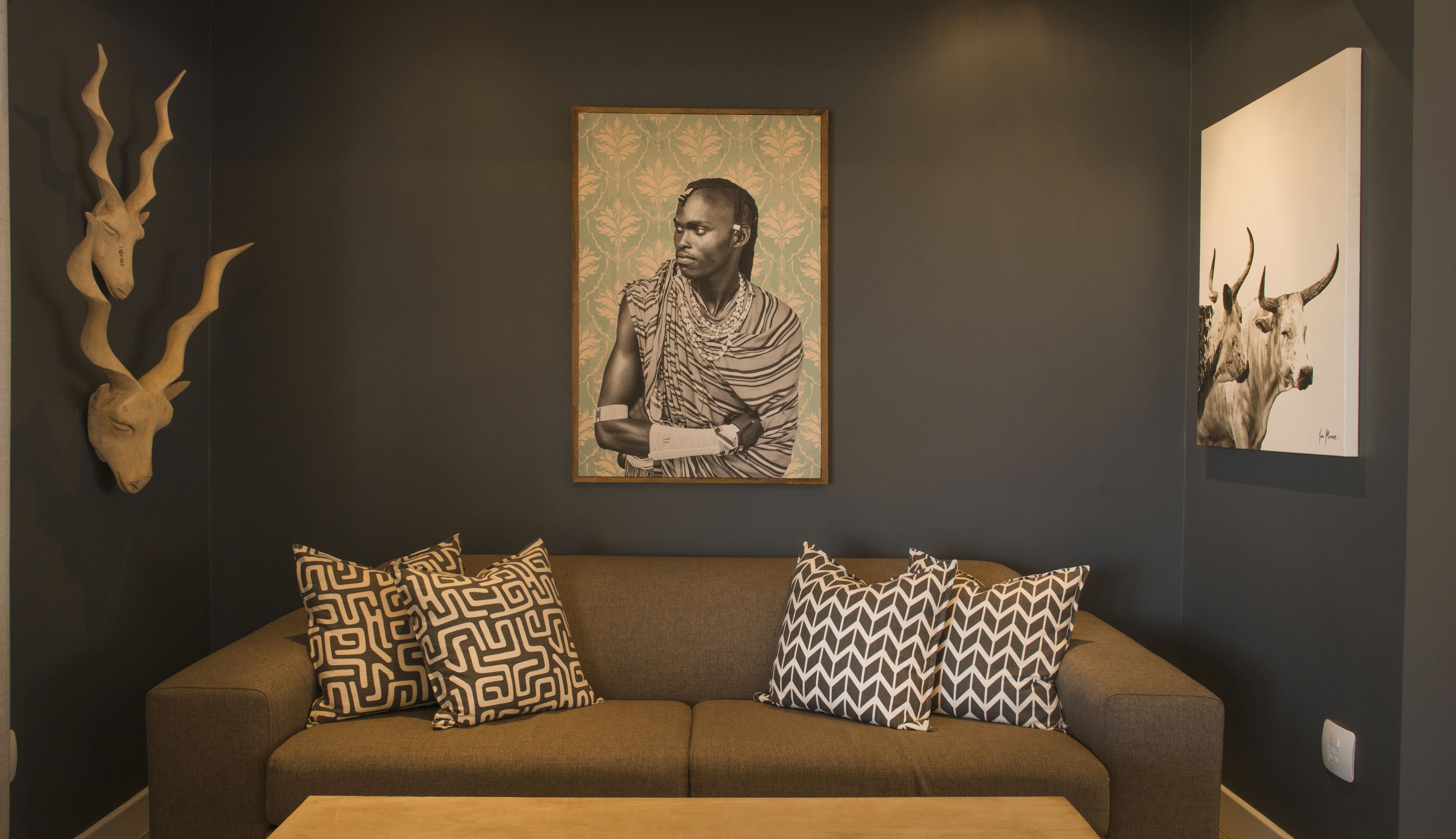 A modern living room with a dark wall, a brown sofa, patterned cushions, a framed artwork of a person, and two mounted animal heads.