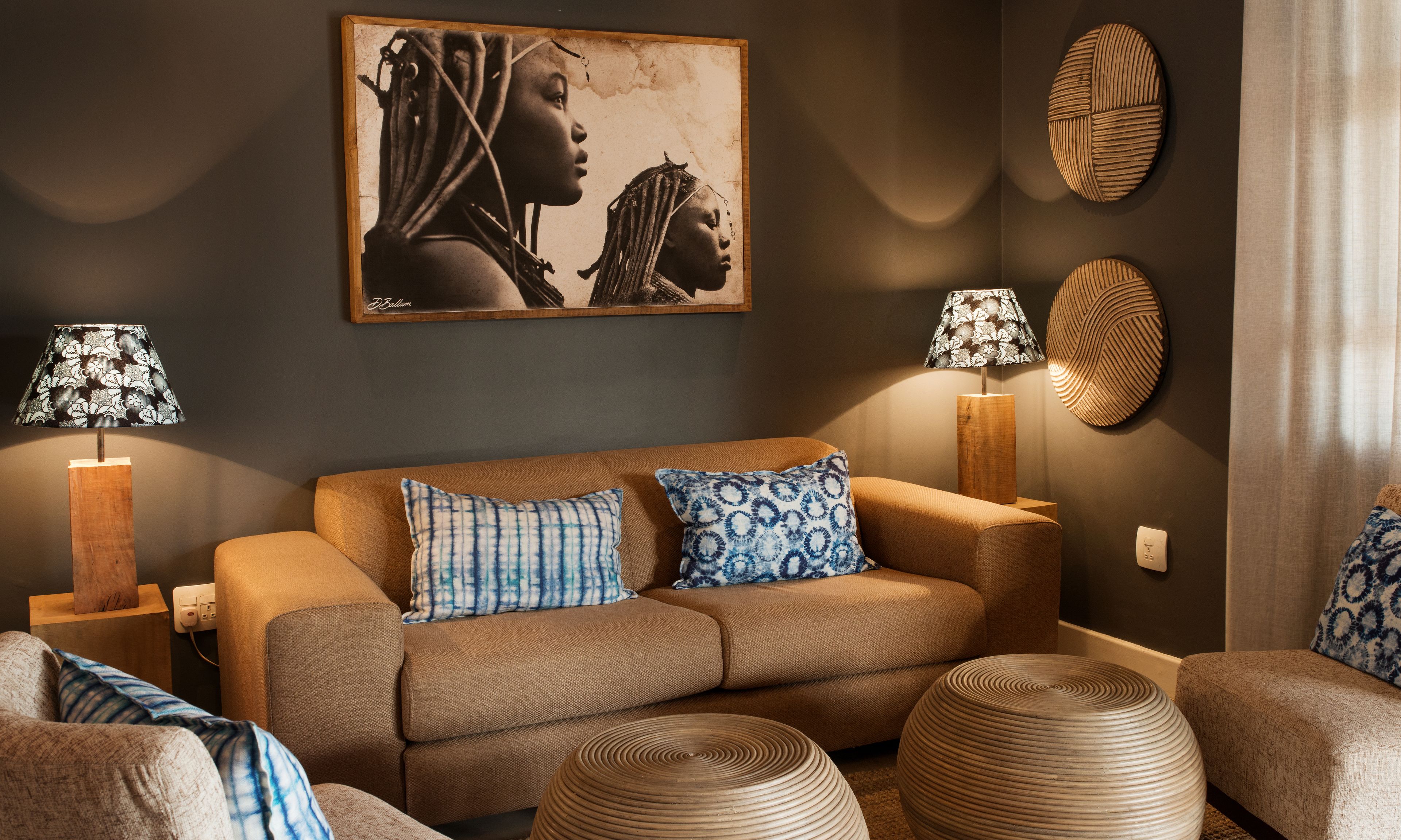 A modern living room with a brown sofa, patterned cushions, two floor lamps, a large framed artwork, and a textured wall decoration.
