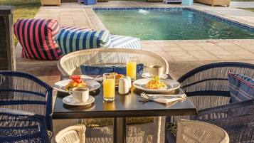 Un área de comedor junto a la piscina con muebles de mimbre, una mesa dispuesta para el desayuno y una piscina al fondo.