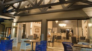 A restaurant with a high ceiling and exposed beams, featuring wicker chairs and tables, and a visible kitchen area. - Greenwood Manor Hotel Ndola (Ndola)
