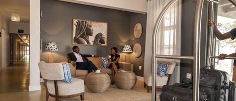 A hotel lobby with a reception desk, a seating area with a sofa and armchairs, a large painting on the wall, and a luggage rack.
