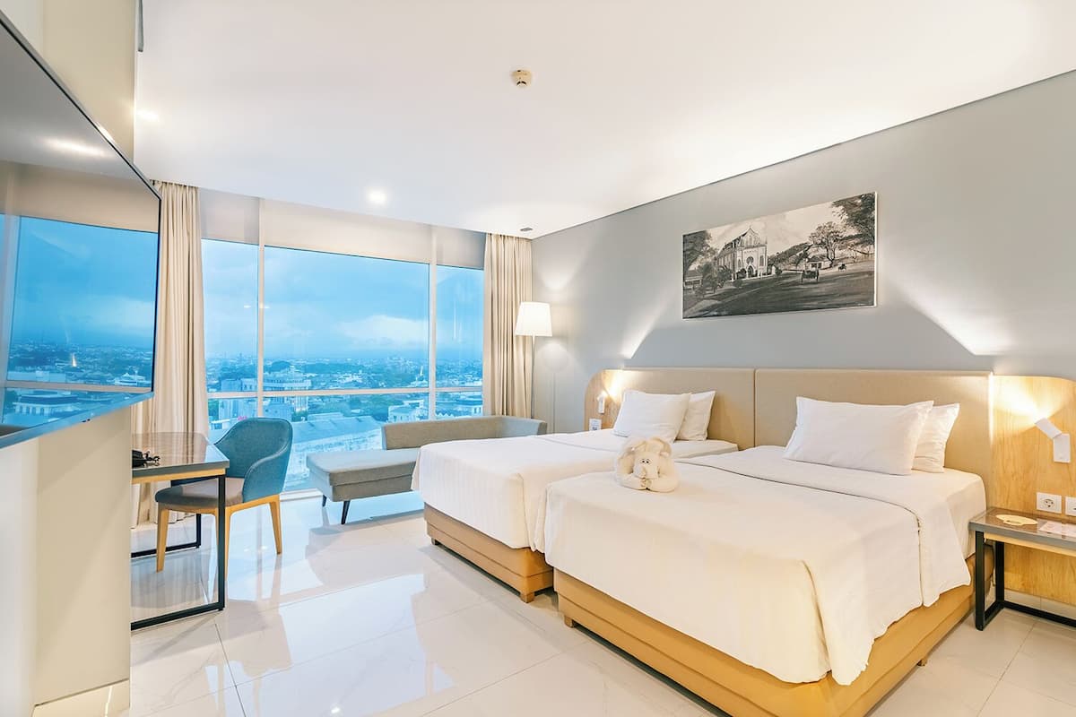 Quarto de hotel com duas camas, uma escrivaninha, uma cadeira e uma janela grande com vista para a cidade.
