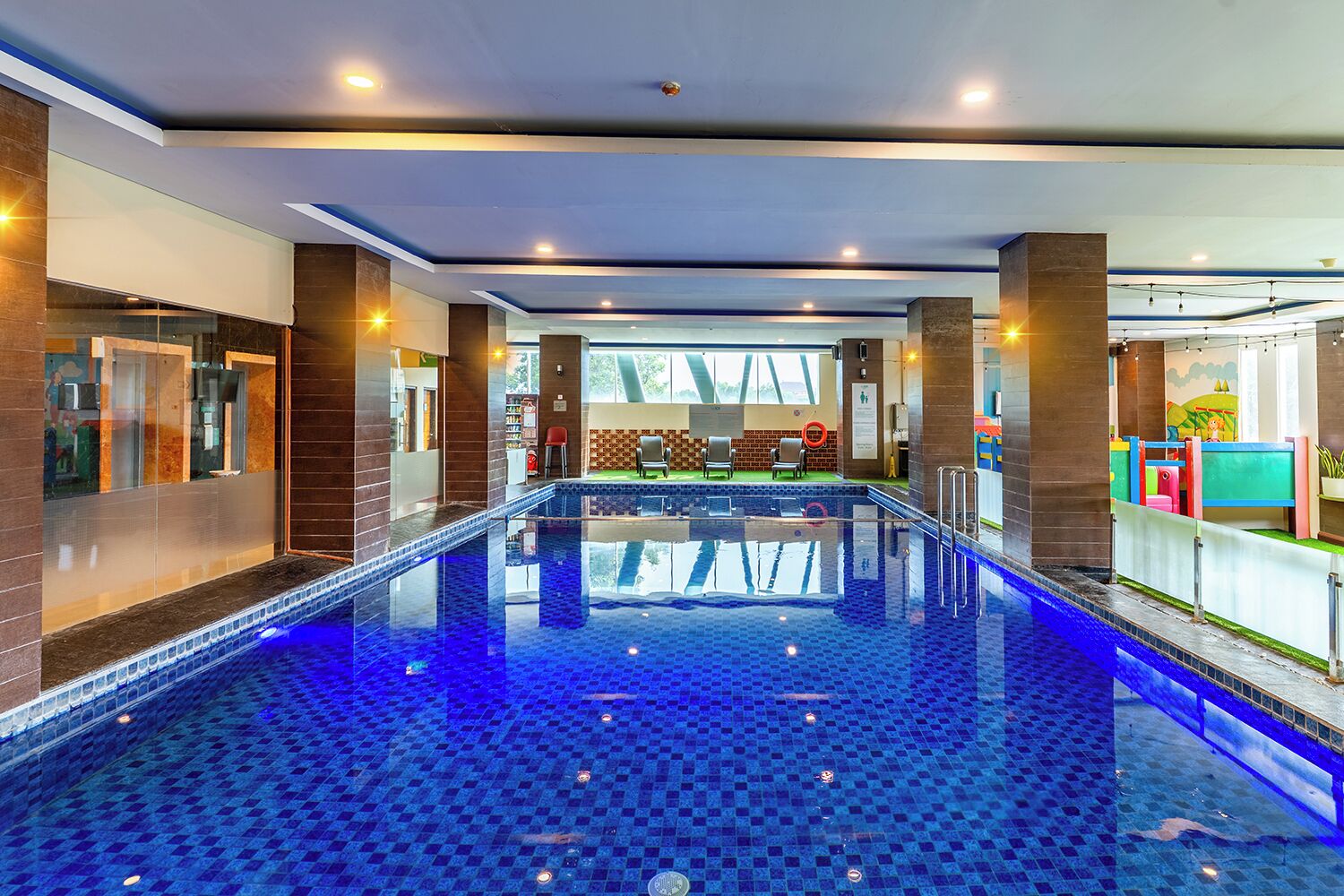 Una piscina interior con suelo de baldosas azules, un área de descanso con sillas y una zona de juegos infantiles con estructuras de colores.