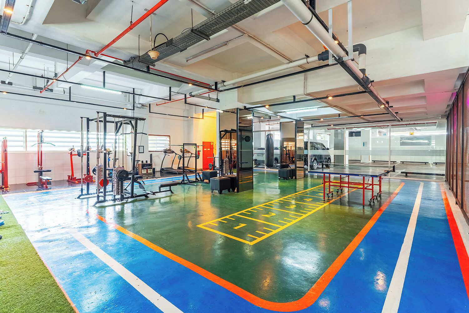 Un gimnasio con equipo de ejercicio, suelo verde y azul, y una zona con rayas rojas y blancas.