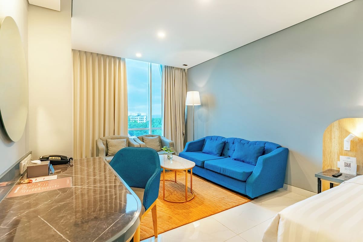 Quarto de hotel com uma janela grande, um sofá azul, uma mesa redonda pequena, uma escrivaninha com uma cadeira e uma cama.