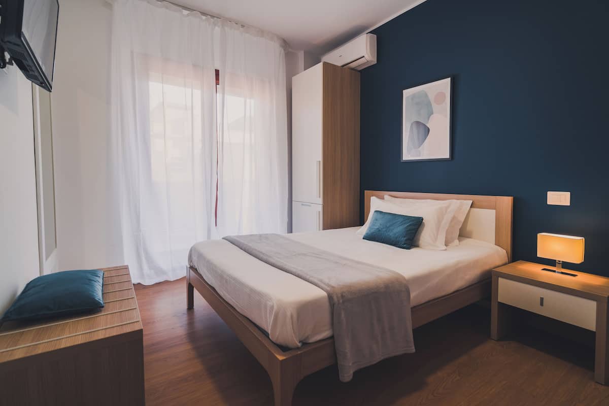 A modern hotel room with a wooden bed, a bedside table with a lamp, a wardrobe, and a large window with sheer curtains.