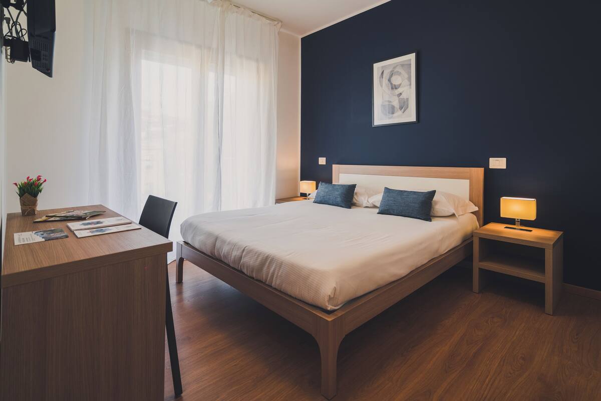 A modern hotel room with a wooden bed, a desk with a chair, a nightstand with a lamp, and a large window with sheer curtains.