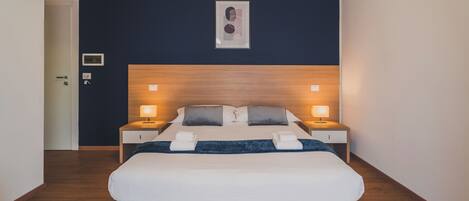 A modern hotel room with a large bed, two bedside tables with lamps, a dark blue wall, and a framed picture above the bed.