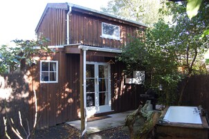 A wooden house with a front door, two windows, and a small garden area with a bench and a tree.