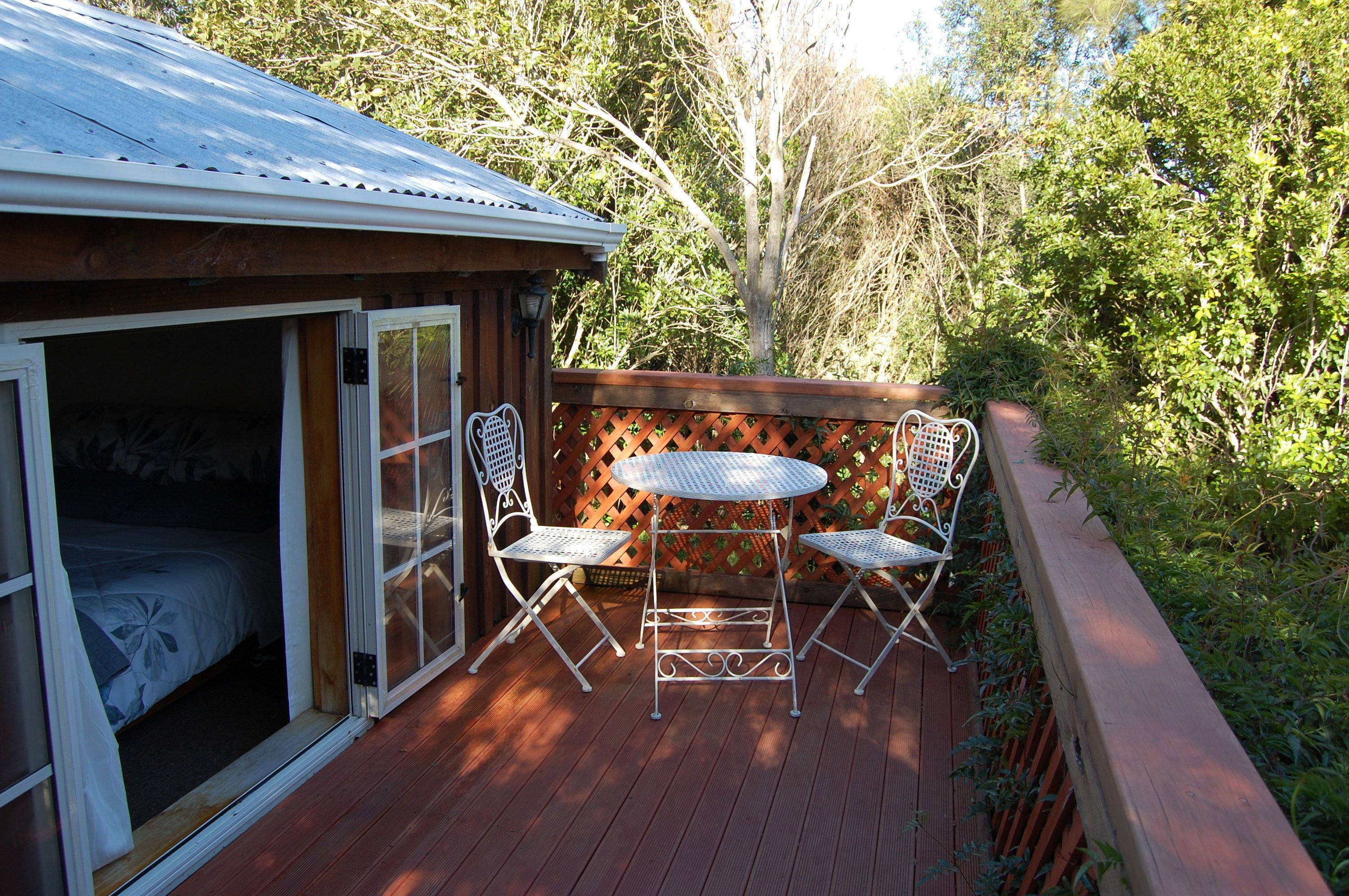 A wooden deck with a metal table and two chairs, a sliding glass door leading to a bedroom, and a wooden fence with a lattice design.