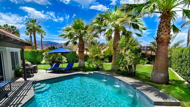 Ein Swimmingpool mit blauen Fliesen, umgeben von Palmen und einer Terrasse mit Liegestühlen.