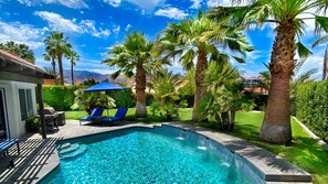 Una piscina con azulejos azules, rodeada de palmeras y un patio con sillones de descanso.