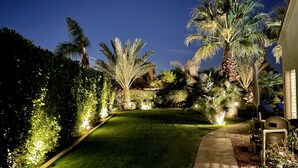 Ein gut beleuchteter Gartenweg mit Palmen und Rasen, der zu einem Gebäude mit einem sichtbaren Fenster führt.