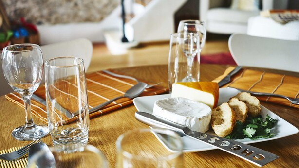 Een eettafel gedekt met kaas, brood en groen, met wijnglazen en bestek.