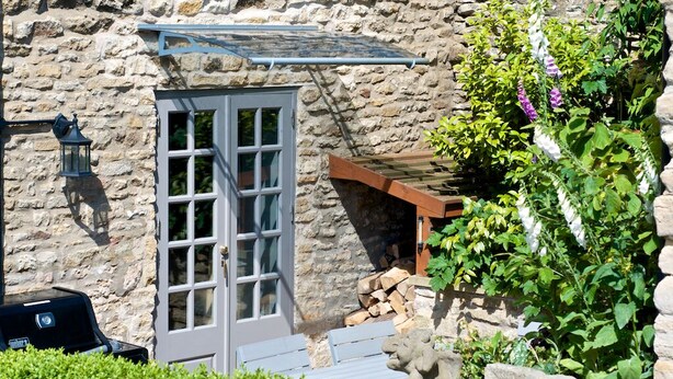 A stone building with a wooden door, a barbecue grill, and a garden with flowers. - Luxuriously Restored Family Friendly 300 year old English Country Cottage  (Westow)