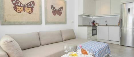 A modern living room with a beige sofa, a round white coffee table with wine glasses and a bottle, and a kitchen in the background with white cabinets and stainless steel appliances.