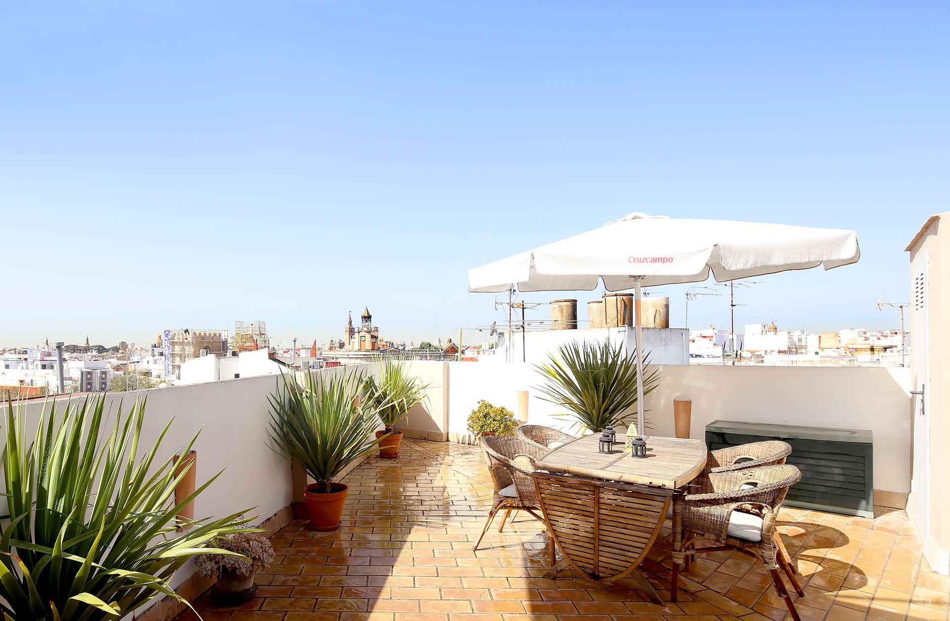 A rooftop terrace with a table and chairs under a white umbrella, potted plants, and a view of the cityscape.