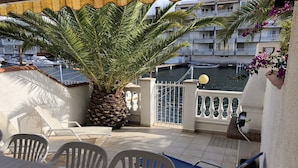 Una terraza con una palmera, sillas blancas, una silla plegable azul y vista al mar y edificios al fondo.