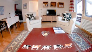 Una sala de estar con dos sillones blancos, una mesa de centro roja con ilustraciones de vacas, un televisor sobre un mueble de madera y una alfombra estampada.