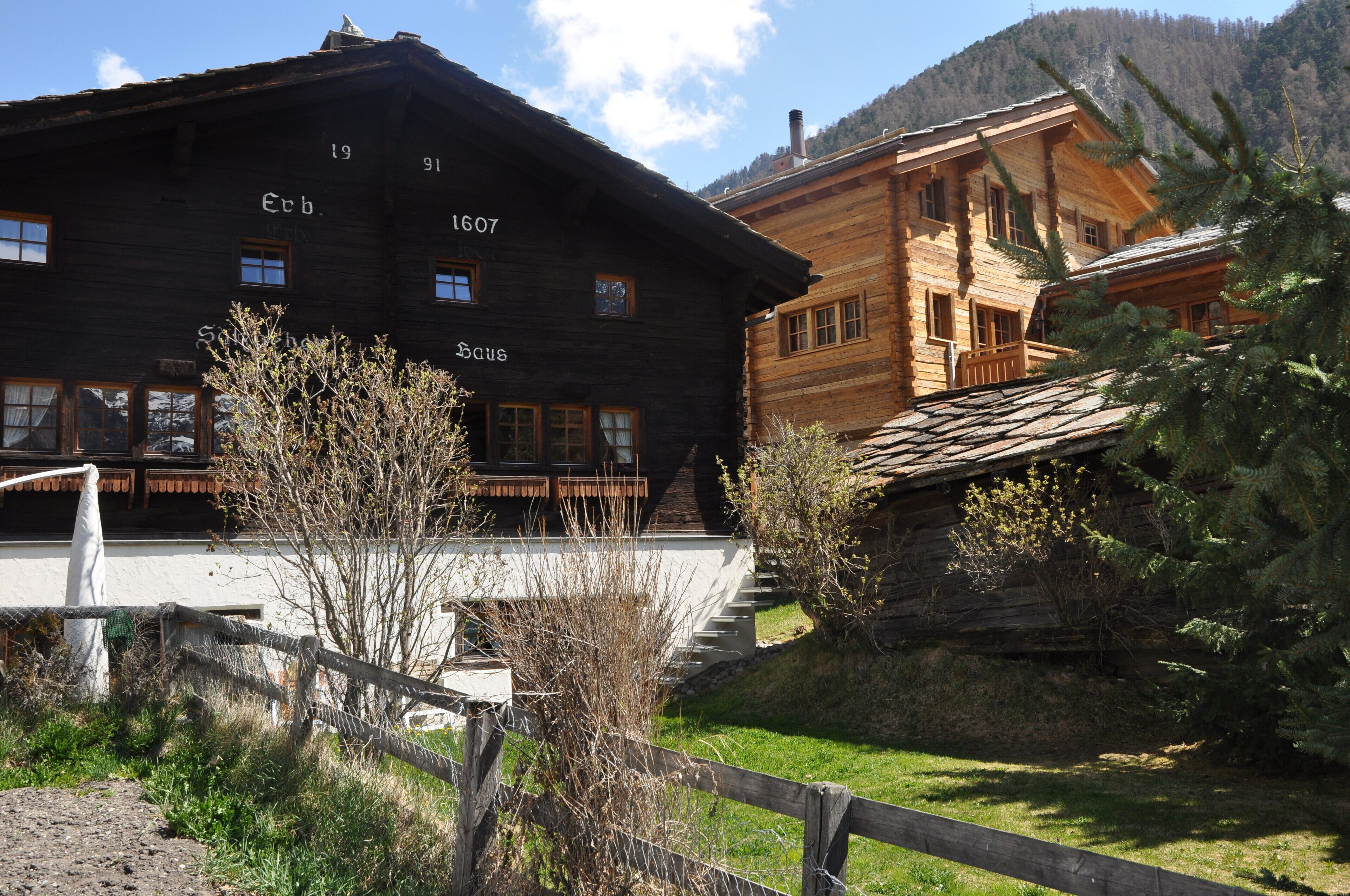Ein traditionelles Holzhaus mit sichtbaren Fenstern und einem Balkon, umgeben von viel Grün und einer Bergkulisse.