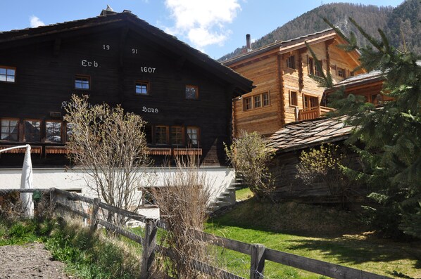 A traditional wooden building with visible windows and a balcony, surrounded by greenery and a mountainous backdrop.