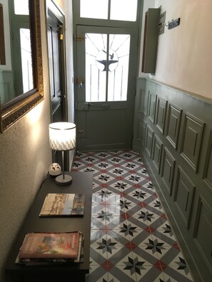 Ein Flur mit fugenreichen Bodenfliesen, einer Tür mit Fenster, einem Wandspiegel und einem Tisch mit Lampe und Büchern.