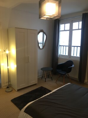 Ein Schlafzimmer mit Bett, Kleiderschrank, Stuhl, kleinem Tisch, Spiegel und einem Fenster mit Vorhängen.
