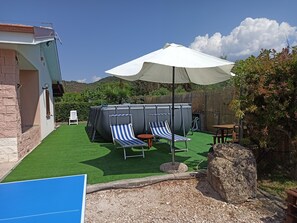 Un espace piscine comprenant deux chaises longues et une table placés sous un auvent, le tout entouré d’une clôture en bois et de verdure.