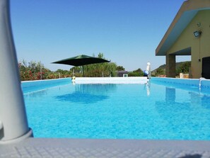 Une piscine d’un bleu éclatant, surmontée d’un parasol vert, entourée de verdure et avec un bâtiment visible au loin.