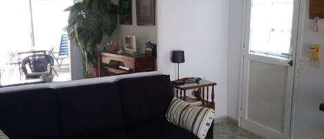 Sala de estar con un sofá oscuro, una mesa de centro con un adorno, una mesita de noche de madera con una lámpara, una planta grande en maceta y una puerta con ventanal.
