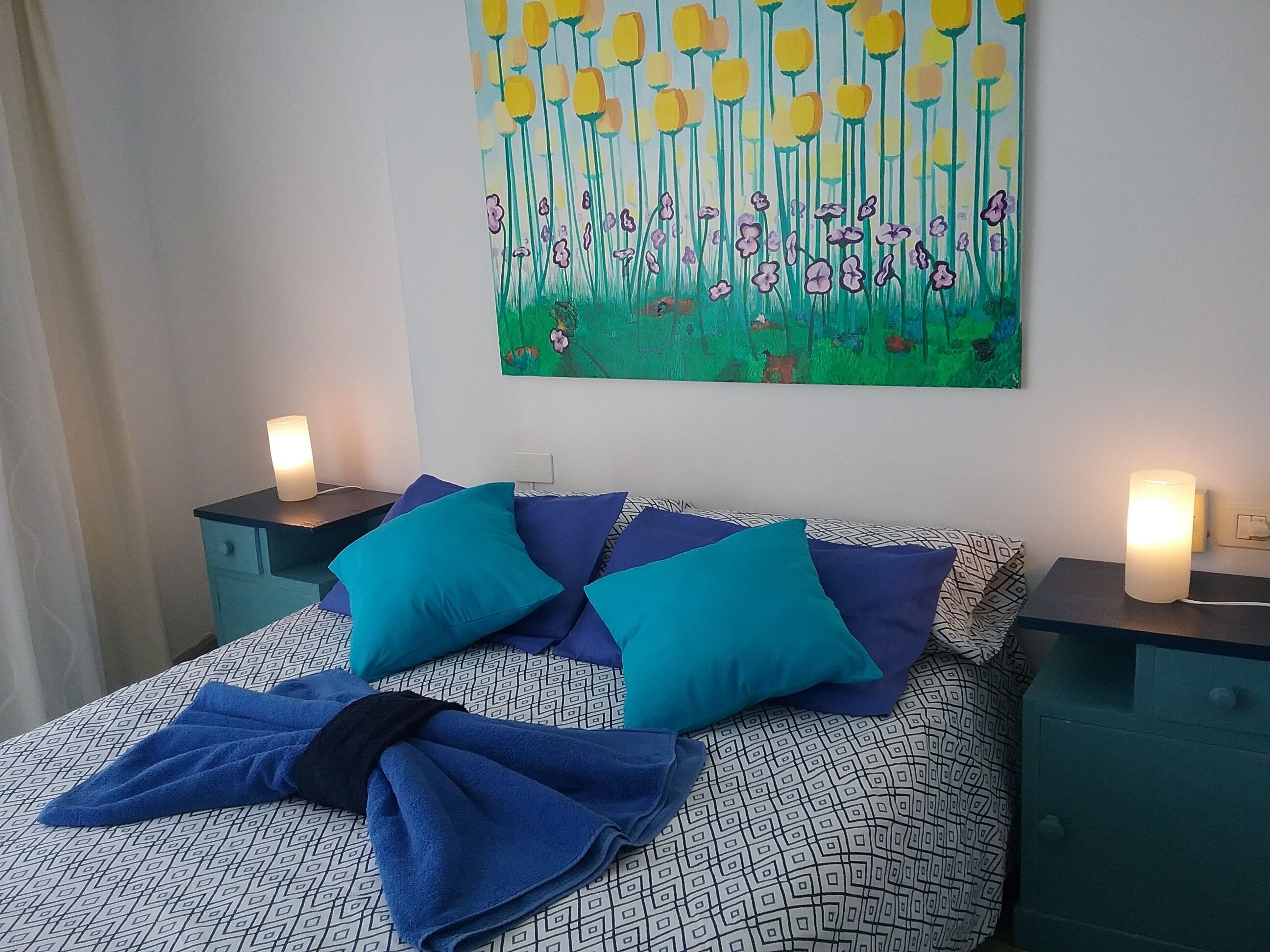 A bed with blue and white patterned bedding, two teal pillows, and a blue towel on the bed. There are two bedside tables with lamps, and a painting of flowers on the wall above the bed.