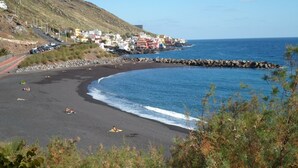Un pueblo costero con una playa de arena negra, casas a lo largo de la costa y un rompeolas rocoso.