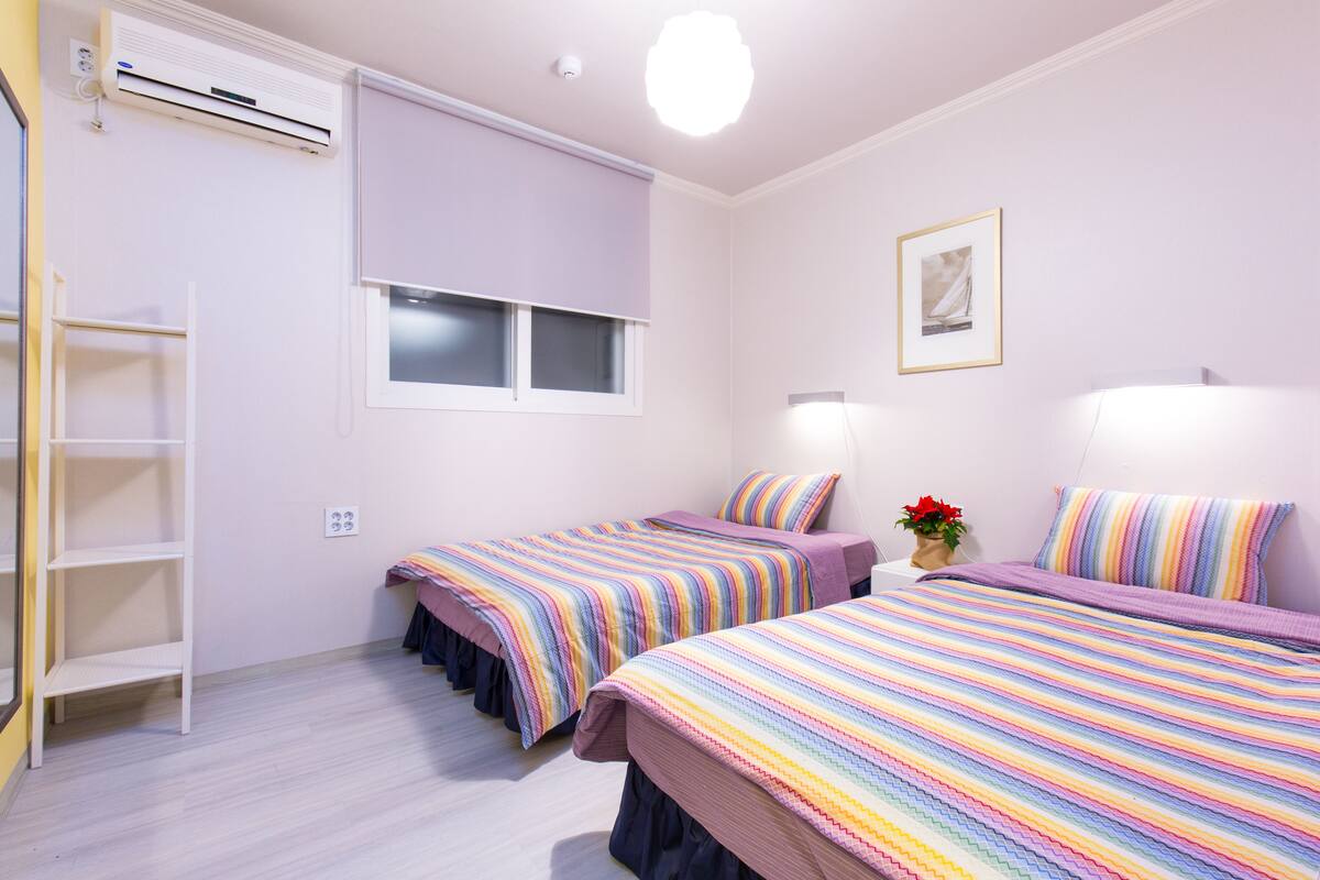 Une chambre avec deux lits, une fenêtre, un appareil de climatisation, une étagère et un tableau encadré au mur.