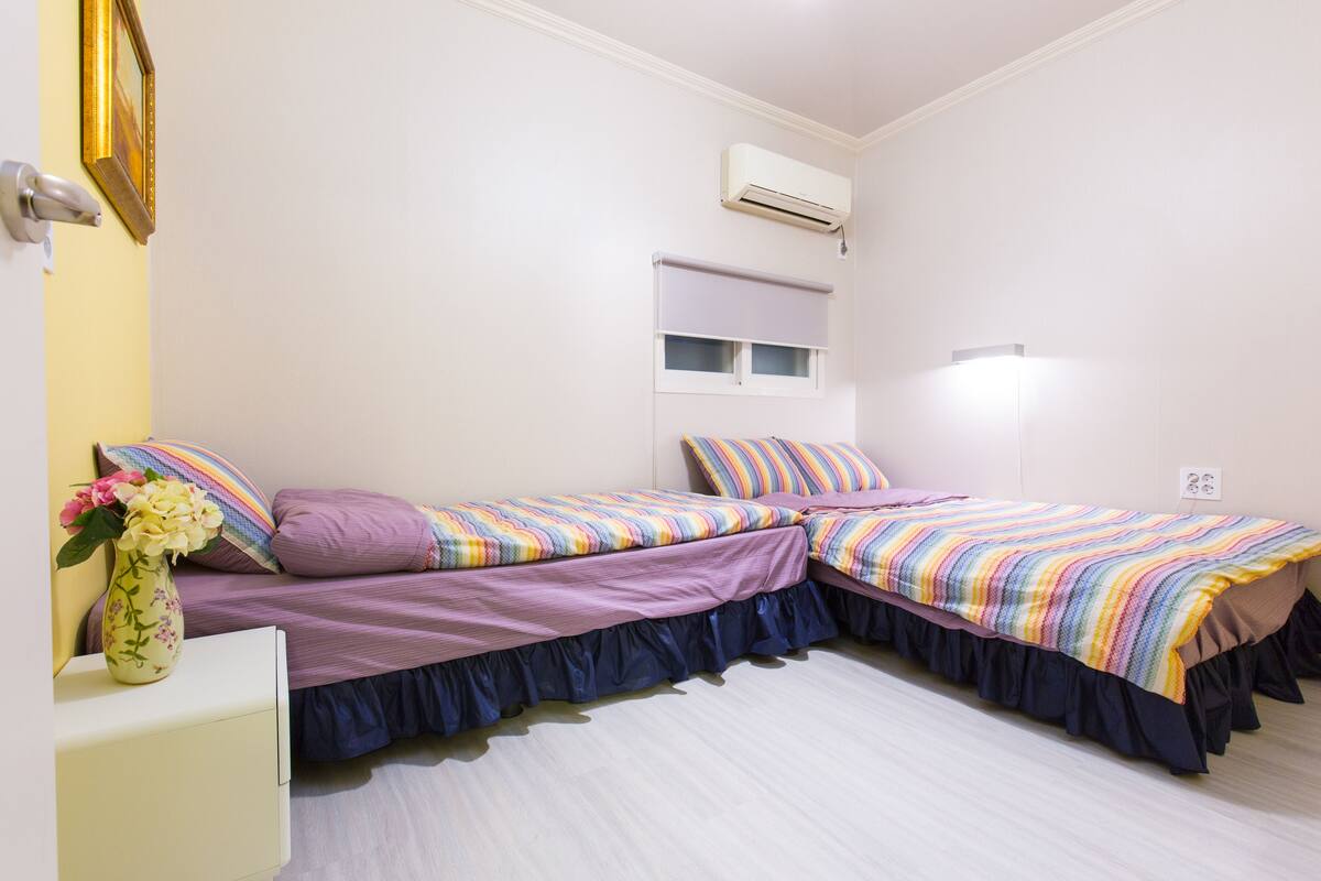 Une chambre avec deux lits, une table de chevet ornée d’un vase de fleurs, un appareil de climatisation et une fenêtre avec des stores.