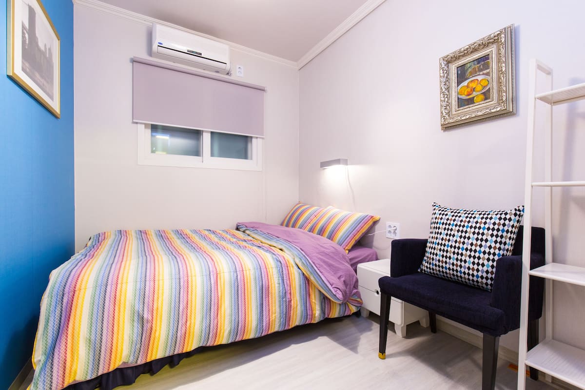 Une chambre à coucher comprenant un lit, une chaise, une table de chevet, un climatiseur, une fenêtre avec des stores et un tableau encadré accroché au mur.
